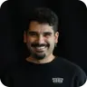 Lächelnder Mann mit dunklem Haar und Bart vor schwarzem Hintergrund.