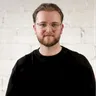 Porträt eines bärtigen Mannes mit Brille und schwarzem Oberteil vor einer hellen Backsteinwand.